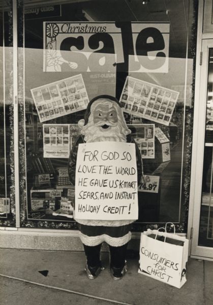 A plastic statue of Santa Claus is standing in front of a store window display advertising a Christmas sale. The statue has a sign hanging around Santa's neck that reads: "For God so love the world he gave us K-Mart, Sears, and instant holiday credit!" On the ground next to him is is a shopping bag with "Consumers for Christ" written on it. This image is a photograph that was glued inside of a Christmas card.