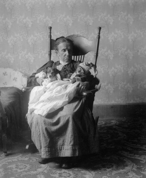 An unidentified woman sits in a rocking chair holding the Middleton family dog, Tootsie, and a cat. The pets are dressed in gowns and the cat wears a sweater and knit hat.