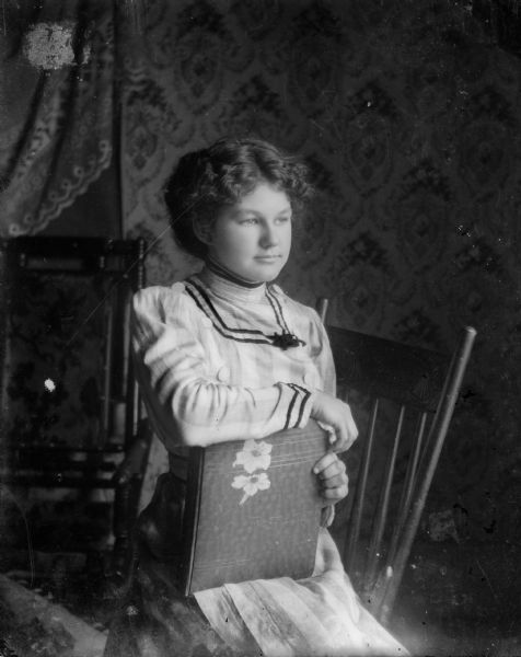 A young woman, possibly Idelle Strelow, poses sitting on a chair and holding a book.