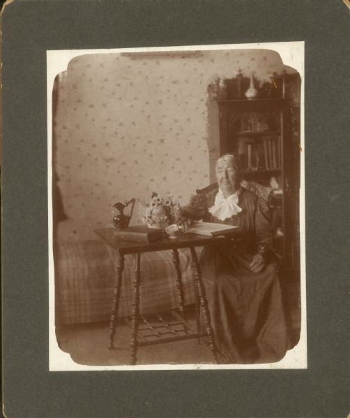 Mrs. Aaron (Joanna) Middleton sits at a table with her hand resting on a book. She was the mother of William Middleton, and mother-in-law of Clara Middleton. A shell, pitcher, bouquet of flowers, and another book also rest on the table. A bed and bookcase are in the background.