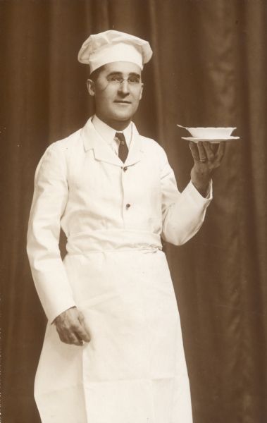 William Middleton, dressed as a chef, poses holding a bowl on a plate.  He is wearing pince-nez glasses.