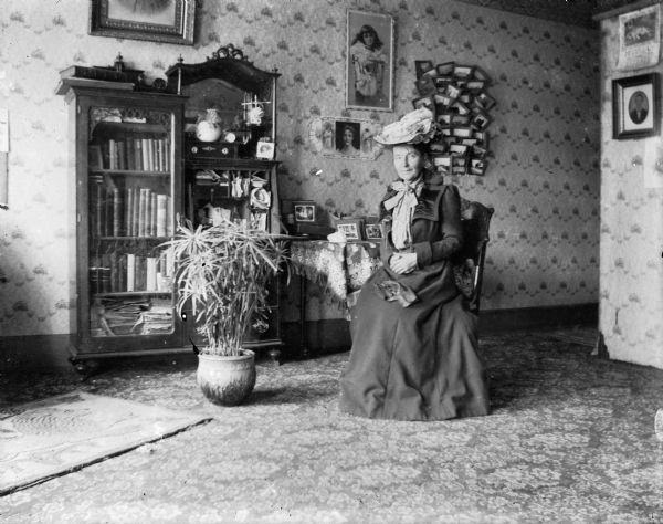 A portrait, possibly a self-portrait, of Clara Middleton seated in her home. She wears a large hat; her gloves rest in her lap. There is a jardiniere with a large papyrus plant in front of a bookcase; prints hang on the wall, and numerous photographs clutter a hanging rack.