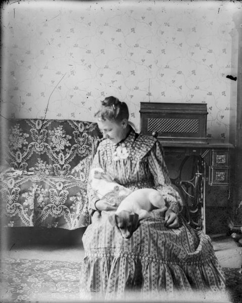 Clara Middleton sits with a dog and rabbit on her lap in what may be a self-portrait. There is a daybed and treadle sewing machine in the background.