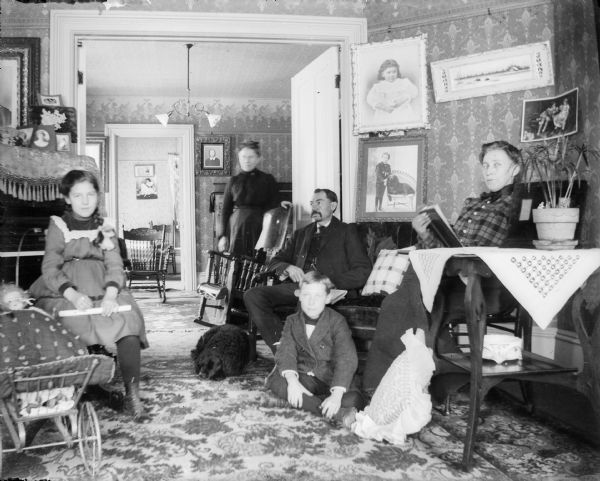 A group portrait of two women, a man, a boy and a girl posing in a parlor; two other rooms are visible through doorways. The girl is seated in front of a piano and next to her is a doll in a toy buggy.  The boy sits on the floor near a curly haired dog. A photographic portrait of a boy and similar dog hangs on the wall behind them.