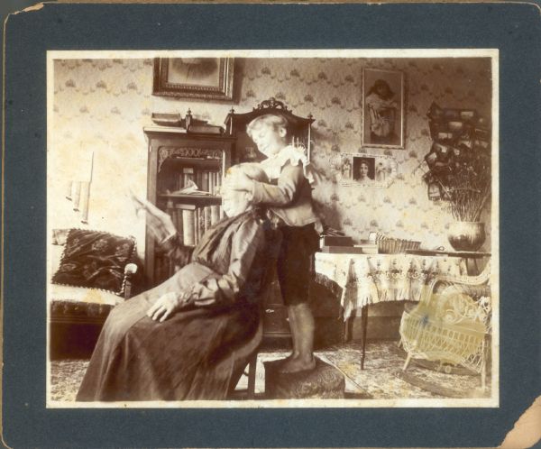 Forest Middleton plays "Guess Who?" with his grandmother. He stands on a small footstool; she is seated in a chair  holding a newspaper or magazine. On the right, there is a rack of small photographs hanging on the wall.