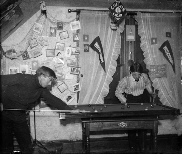 Forest Middleton, left, prepares a pool shot while his opponent looks on. They are playing on a portable pool table in Forest's room. Photographs, cigar advertisements, and two Wisconsin pennants decorate the room.