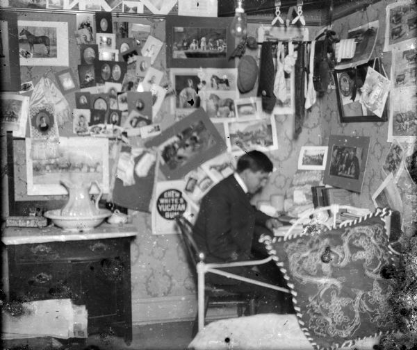 Forest Middleton reads at his desk in a room cluttered with photographs, neckties, suspenders, and a cap. A bare lightbulb hangs from the ceiling. There is a commode with china commode set and an iron bed.