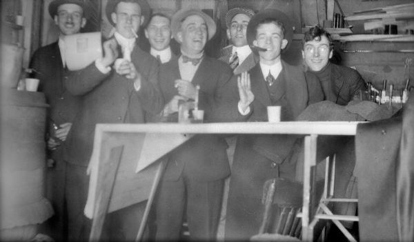 Forest Middleton, far right, poses with a group of men in high spirits. One man, second from left, holds a jug. The room is furnished with a makeshift table and woodburning stove.