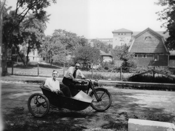 Forest Middleton poses in a sidecar smoking a pipe; the driver of the motorcycle is unidentified. They are parked on West Johnson Street with the University of Wisconsin-Madison's Lathrop Hall in the background; a house and barn sit between Johnson Street and University Avenue.