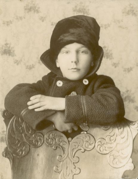 Forest Middleton rests his arms on a press-back chair. He wears a hat and winter coat.