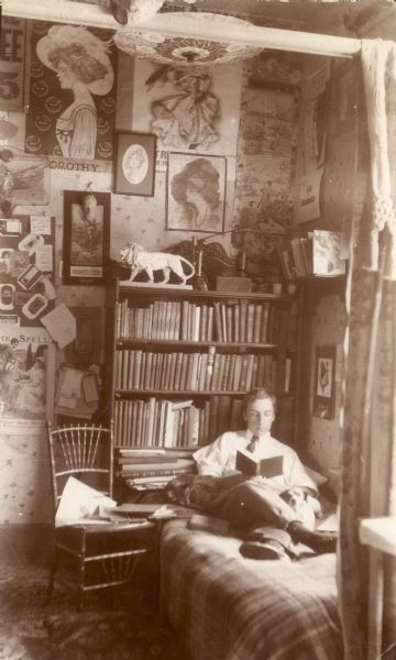 Photographic postcard of Forest Middleton reading while lying on his bed with the family dog, Tootsie, beside him. There is a full bookcase behind the bed, and a chair alongside. The room is decorated with posters, prints, a paper parasol, and a statuette of a lion.