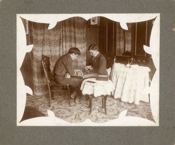 Forest Middleton, left, and Arthur Strelow, play chess with the board resting on their knees. Drapes hang in a doorway; there are also two round tables with tablecloths in the room.
