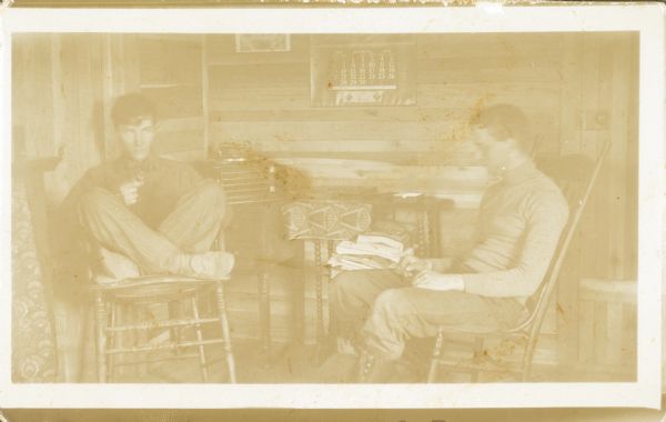 Photographic postcard of Forest Middleton, left, sitting cross-legged on a chair holding a pipe and glancing at a friend, possibly Eddie Quick. There is a phonograph in the background and a 1913 calendar on the wall. The room is paneled with wood.