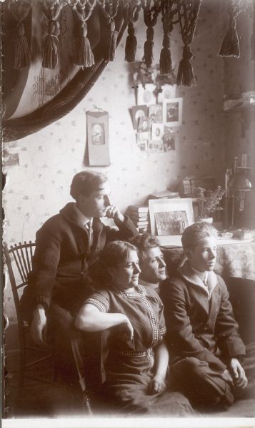 Photographic postcard of Forest Middleton sitting on a chair behind an unidentified man, woman, and boy. A heavy drape with tassels, top left, frames the scene. There is a rack of photographs on the wall; a book, lamp and photograph rest on a table behind the group.