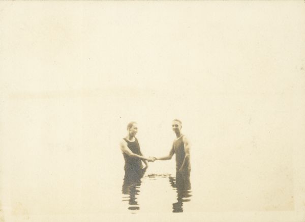 Two men in swimming suits shake hands while standing in thigh deep water.