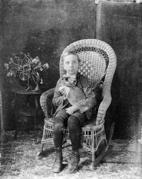 Forest Middleton sits in a wicker rocking chair holding a rabbit. There is a plant on a small stand on the left.