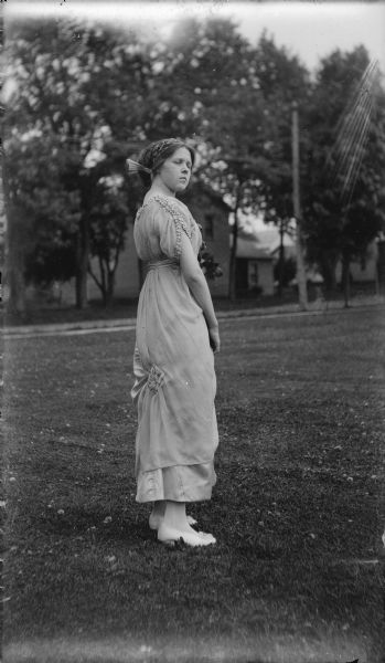 Leonore Judkins, later Mrs. Forest Middleton, poses standing outdoors in a fancy dress, with flowers at her waist. She is wearing her hair in a bun and wears a headband with a fan-shaped embellishment.