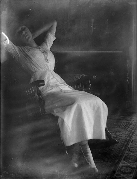 Leonore Judkins, later Mrs. Forest Middleton, poses in a rocking chair with her hands behind her head. She wears a fancy dress and high buttoned shoes. There is an upright piano in the background and an Oriental carpet on the floor.