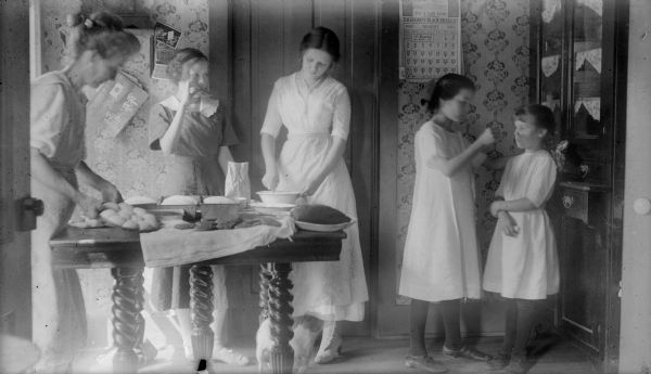 Leonore Judkins, later Mrs. Forest Middleton, center, prepares baked goods with her mother, far left, and sisters, Juanita, Bonnie, and Blanch. Bread and rolls are rising on the table, where there is also a mound of cookies. A calendar on the wall advertises Thedford's Black-Draught, "For a Lazy Liver." There is a "ghost" image of Tootsie, the Middleton family dog, under the table. 