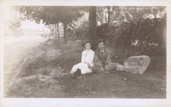 Forest Middleton and his future wife, Leonore Judkins, sit on the ground against a tree alongside a country road. Grapevines grow on a fence on the right.