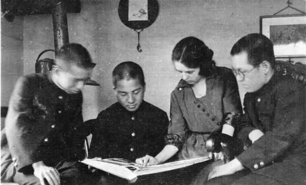 Leonore Judkins Middleton showing a book to three young Japanese men. An iron kettle is sitting on a stove in the background. On the reverse of the print is written: "Taken at Hagin's one Sun. a.m. I am showing our album of pictures." Fred E. Hagin was a missionary of the Disciples of Christ Church in Japan.