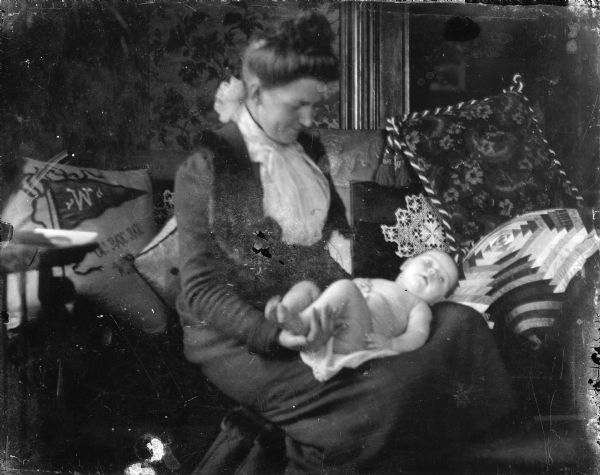 Clara Middleton holds her naked infant son Forest on her lap. There are many throw pillows around them, including one that features a "W" and the cheer "U! RAH RAH" on it.