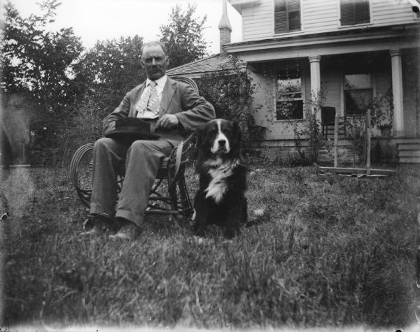 A man sits in a bentwood chair, holding his hat in his lap, as his dog sits nearby. They are sitting on the front lawn of a large house.