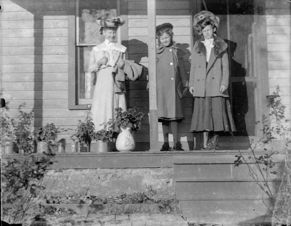 A woman and two girls stand on a porch wearing large hats. Potted plants line the porch.