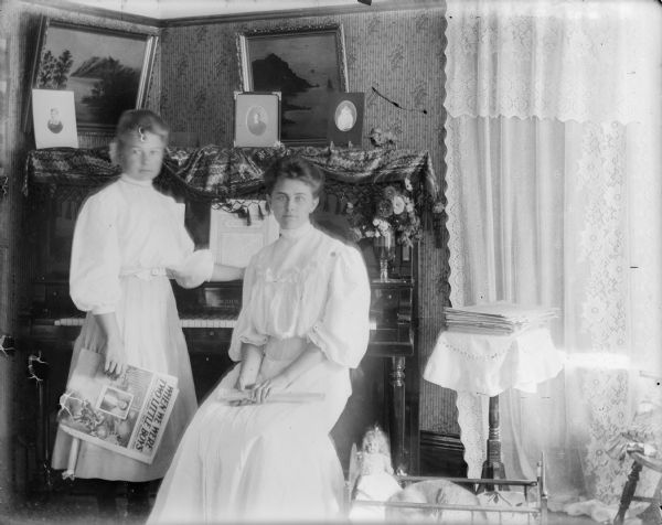 Two well-dressed young women pose in front of a piano. One holds a fan in her lap, the other holds a piece of sheet music titled "When We Were Two Little Boys." The room is lavishly decorated with paintings, lace curtains, piano scarf, and flowers in a vase on the piano. A doll and sleeping cat occupy a doll bed on the floor.