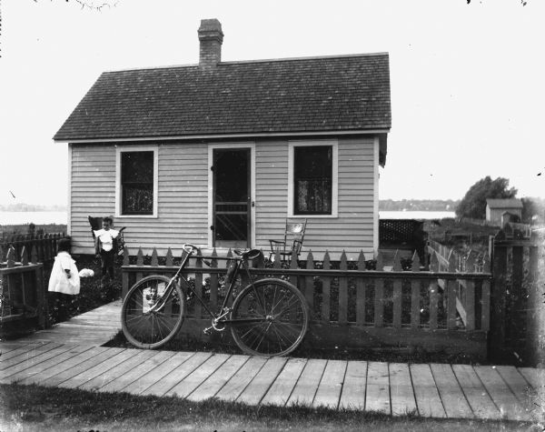 View from road of two children standing in the yard of a small cottage on a lake. There is a white rabbit on the ground between them. A bicycle leans against a picket fence; a board sidewalk leads to the front door. There are two chairs on the lawn.