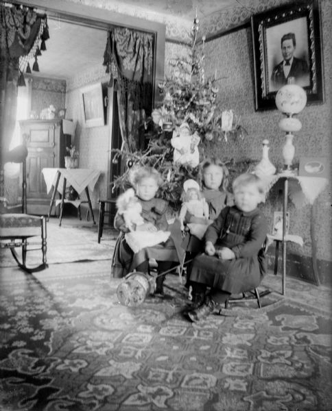 Three unidentified children sit in chairs in front of a Christmas tree. Two of the children hold dolls; the third holds a push toy. The tree is decorated with tinsel, glass ornaments, candles, and large Santa and angel.