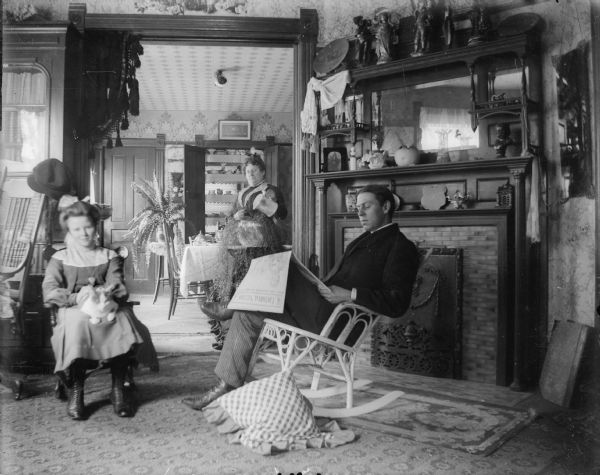 A carefully posed scene of a man, woman, and girl at home. The girl sits with a cat on her lap. The man, in a wicker rocker, reads the "Editorial Section" of a newspaper; the woman waters a fern. The doors of a built-in cabinet in the background have been opened to reveal decorative china.  On the right is a fireplace with ornate mantel displaying bric-a-brac. A heavy drapery with tassels decorates the doorway between the parlor and dining room.