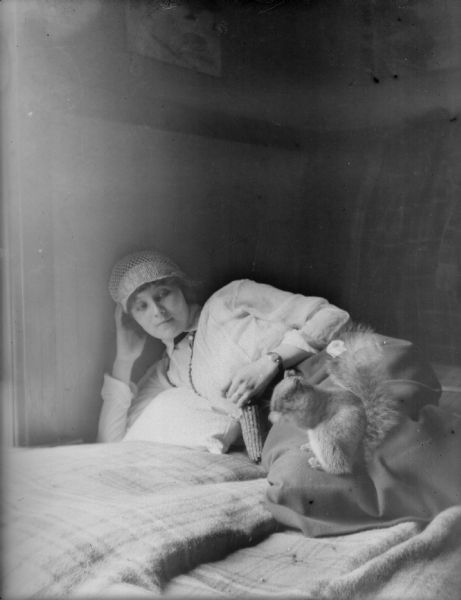 A young woman holding an ear of corn reclines on a bed while a squirrel sits on her thigh. The woman wears a heavy skirt, blouse, and needlework cloche hat.