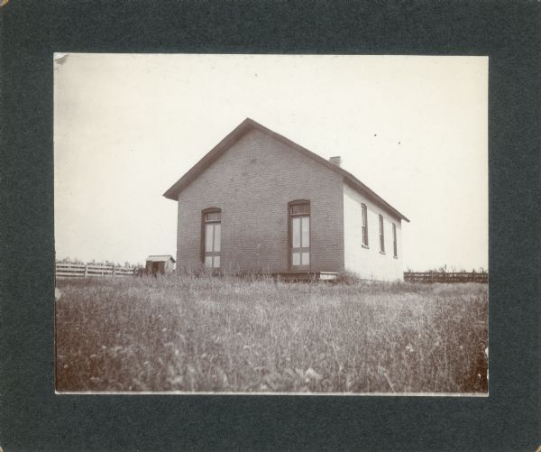 A brick, one-story country schoolhouse with two entrances, and an outhouse on the left. The schoolyard is fenced.