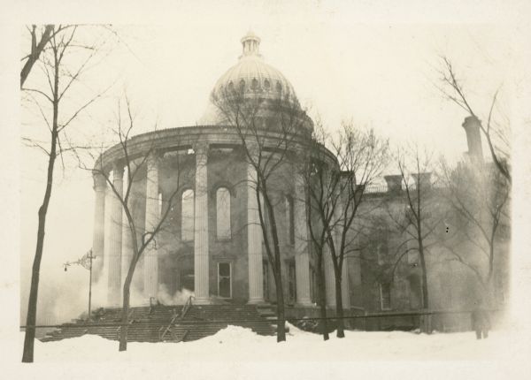 A view of the smoldering third Wisconsin State Capitol. The roof of the west wing has collapsed; debris litters the steps.