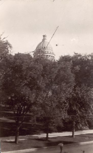 A crane lowers the flag pole from the third Wisconsin State Capitol as the first step in demolition of the dome. An iron eagle is perched atop the flag pole and is at the right end of the pole. It is now on display at the Wisconsin Historical Society headquarters building. The capitol building suffered severe damage in a fire in 1904. Demolition was carried out in stages, coordinated with the building of the new capitol.