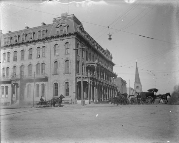 A view of the Second Empire style Park Hotel at the corner of South Carroll and West Main Streets. There are horse-drawn carriages, pedestrians, and a small dog on the streets. Grace Episcopal Church is visible in the background; the Capitol Park is on the right. Overhead are electric wires for streetcars and a light.