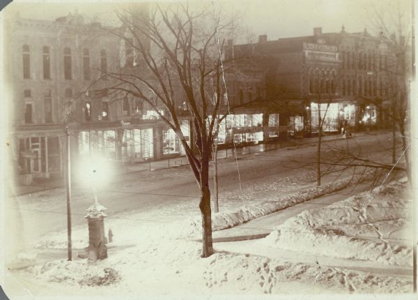 An elevated night view of North Pinckney Street and the north corner of Capitol Park. Walzinger's Drugstore and the Olson and Veerhusen clothing store signs are visible. There is snow on the ground.