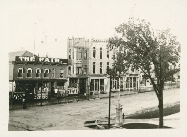 Elevated view of the businesses on the east side of North Pinckney Street, including The Fair, The Madison School of Music, and August Scheibel. In the foreground, pillars with lamps stand on either side of the sidewalk into the Capitol Park.