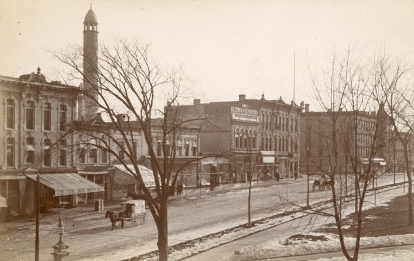 Elevated view of horses and buggies traveling on North Pinckney Street. The water tower rises behind the commercial buildings on the east side of the street. Walzinger Drugstore is identified on its awning. Olson and Veerhusen's "The Big Store" is also identified. The Capitol Park is in the foreground.