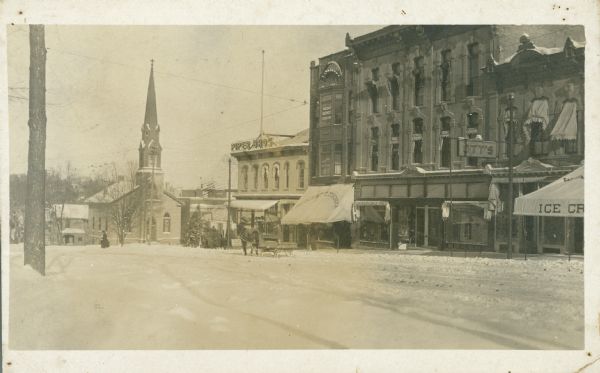 A view of the Evangelical Society's Immanuel Church on Hamilton Street, as seen from North Pinckney Street. Snow covers the ground and a horse-drawn sleigh is seen. The commercial buildings along Pinckney Street include Piper Bros.; Madison Business College and Crescent Clothing; Wilkinson Daly Hats; Ott's; and Walzinger's Drugstore which advertises ice cream on its awning.