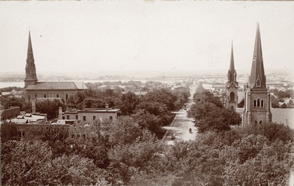 Elevated view down West Washington Avenue from the roof of the Wisconsin State Capitol. Three churches are seen, from left: St. Raphael Catholic Church, First Congregational Church, and Grace Episcopal Church. Lake Monona is on the left. The trees of Capitol Park form the foreground.