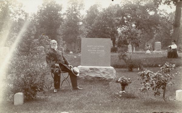 Captain William Hogbin sits on a chair next to the headstone of his late wife, Mary Catherine (Tiffany Knott) Hogbin, in Forest Hill Cemetery. A woman, likely his second wife, sits in the background.  Captain Hogbin was born in England and trained as a tailor there. He immigrated to the United States with his parents and served in the Civil War in Company F, Eighty-eighth Illinois Volunteer Infantry. He was active in the Lucius Fairchild Post of the G.A.R. and had a tailor shop in Madison for many years.