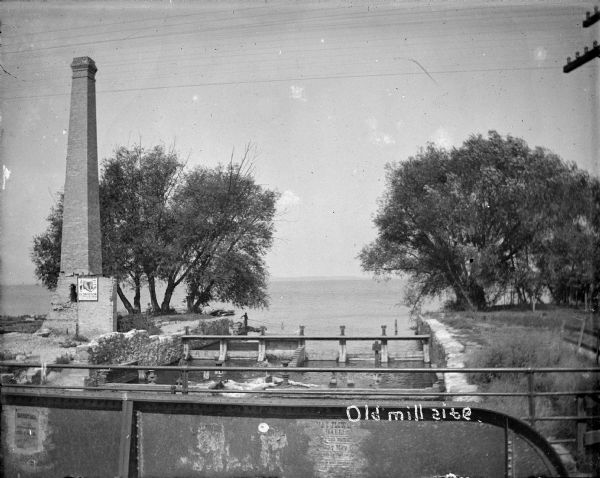 View from bridge towards a tall smokestack standing on the left side of the Yahara River where it is flowing out of Lake Mendota. Remnants of the foundation and supports of Farwell's Madison Mills are seen in and along the river. A man is fishing from the shore.