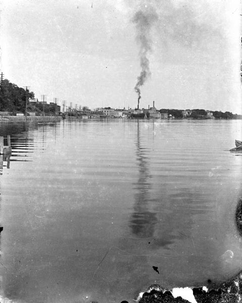 A view east along the north shore of Lake Monona, with Madison's east side industrial area in the background. The Deering Harvester Company is identified by a sign painted on its wall. The tallest tower right of the smokestack is part of the Fauerbach Brewery complex. The short domed structure on the shoreline is a gazebo which was also a part of the Fauerbach complex. Large private homes are seen on the far right. Tall utility poles are along the shoreline and railroad tracks.