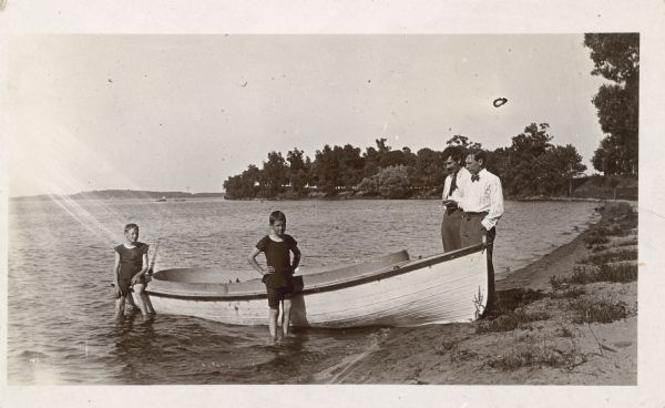 Two boys in swimsuits and two well-dressed men pose beside a rowboat which has been pulled up on the sandy shore of, most likely, Lake Mendota.