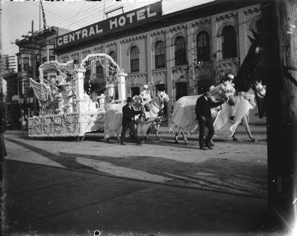 A parade float elaborately decorated with flowers and pulled by four robed horses is traveling west along East Doty Street, with the Central (Fess) Hotel in the background. A woman is riding on the float in a winged throne. Two men in white are riding horses, while two other men in street clothes are walking alongside. A dark horse's head behind a utility pole is in the right foreground.