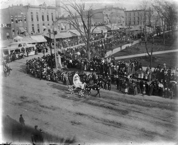 Elevated view of a crowd watching a parade at the corner of North Pinckney and East Mifflin Streets. There is a temporary viewing platform set up in front of "The Fair" store on Pinckney Street. A black horse pulls a carriage carrying two female figures which appear to be mannequins. Both horse and carriage are decorated with large chrysanthemums.