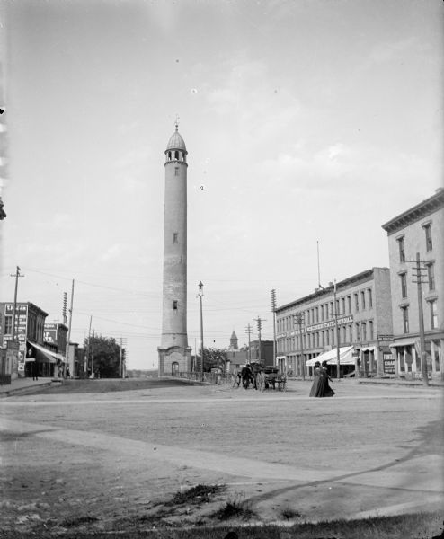 The 125 foot tall Madison water tower stands in the middle of East Washington Avenue. On the right is a large business block occupied by the Amerika Publishing Company, Grimm Book Bindery and the "Wisconsin State Journal." Two women are walking across the street; a man is standing beside a horse-drawn wagon.
