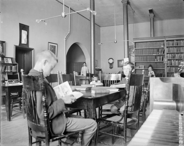 A view of the interior in the new Madison Public Library when it was located in the city hall, West Mifflin Street. Two boys read at a table in the foreground. Children and two librarians are in the background. Bookcases line the back wall. A piano keyboard is in the right foreground. Light fixtures with bare bulbs hang from the ceiling.